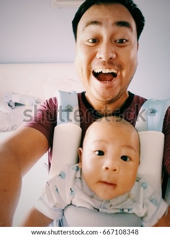 Shot of happy father while taking pictures of his Asian baby boy with a smartphone at home. Dad and kid smiling while taking selfies together in the bedroom. Family concept