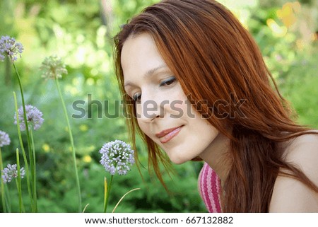 A beautiful young girl with long red hair looks thoughtfully at the sky against a background of summer green foliage and lilac flowers