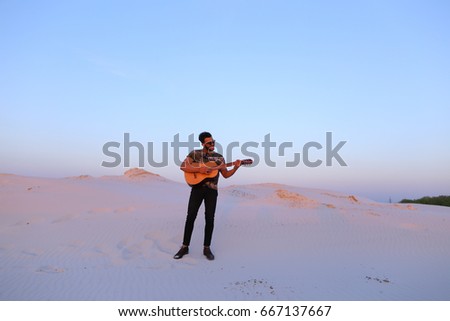 Young Muslim man stands at full length and plays melody on stringed musical instrument, takes chords, smiles and sings to himself, inspired by surrounding landscape, standing in midst of wide sandy