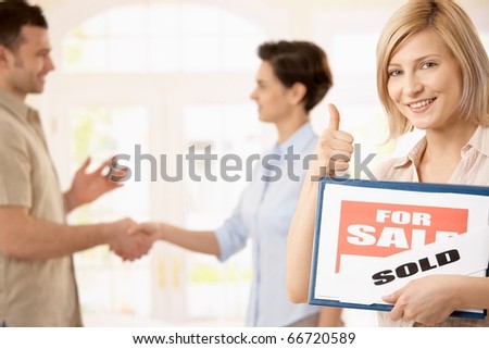 Happy woman holding for sale sign giving the thumb up, smiling man shaking hands with estate agent in background.?