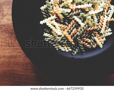 It's time for Italian menu. The picture of multicolored of dried fusilli consist of yellow, orange and green on black plate above wooden table.  soft tone color