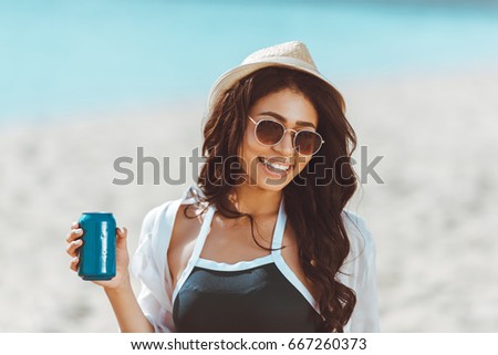 Beautiful young woman in sunglasses and straw hat holding soda can and smiling at camera  Royalty-Free Stock Photo #667260373