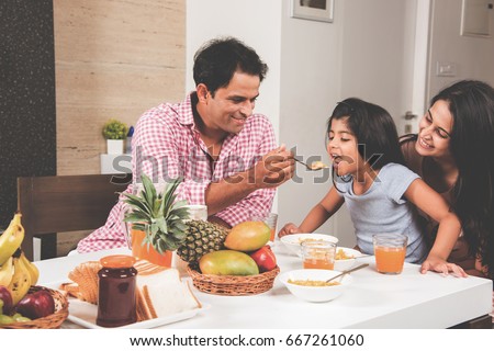 An attractive happy, smiling Asian Indian family of mother, father and daughter. Father feeding cereal to daughter with spoon at dining table. Indians eating breakfast, lunch / dinner. Selective focus Royalty-Free Stock Photo #667261060