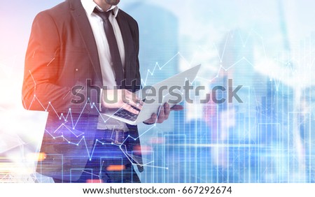 Side view of a young bearded businessman holding a laptop and standing against a modern cityscape with graphs in the foreground. Mock up toned image double exposure