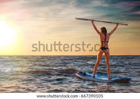 Woman practicing SUP yoga at sunset, meditating on a paddle board on sunset.
