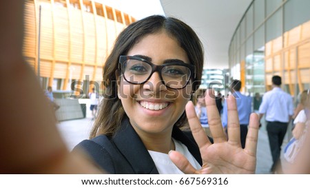 Portrait of a young beautiful business woman (student) in a suit, glasses, walking through the city, video call, selfie, holding a phone. Concept: new business, communication, Arab, banker, manager. Royalty-Free Stock Photo #667569316