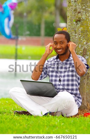 Young black man sitting down on green grass and working in his computer and listening music with his headphones posing his back in a tree in the city of Quito Ecuador