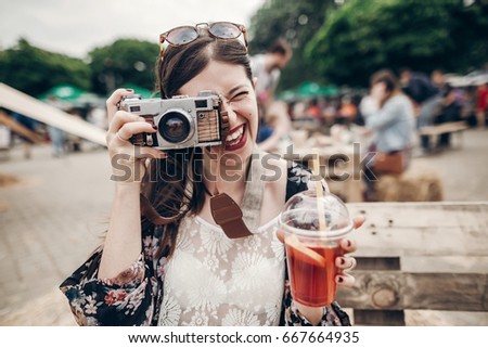 happy hipster woman in sunglasses making photo with old camera and drinking lemonade. stylish boho girl holding cocktail and smiling at street food festival. summertime 