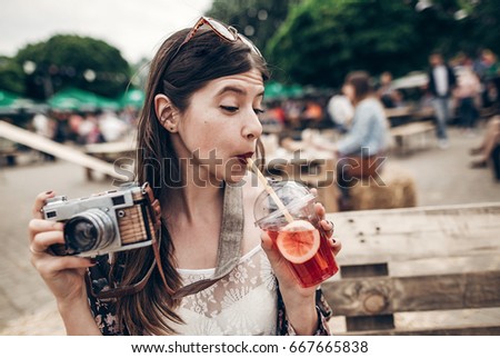 stylish hipster woman in sunglasses with red lips drinking lemonade and holding old photo camera. boho girl holding cocktail and smiling at street food festival. summer  travel 