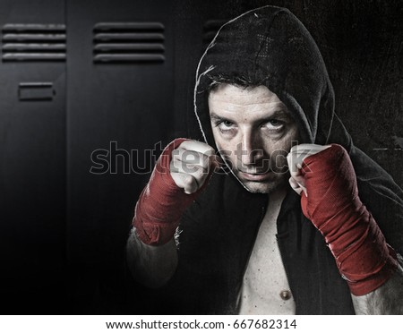young man in boxing hoodie jumper with hood on head wearing hand and wrist wrapped ready for fighting posing isolated on gym locker room grunge dirty background with angry face expression