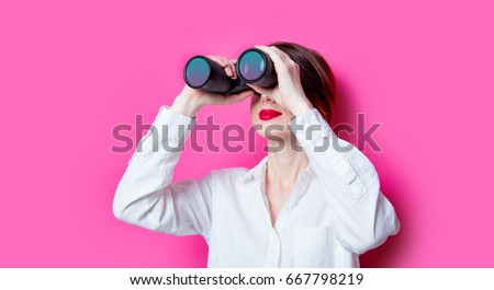 Beautiful red-haired businesswoman with binoculars on pink background