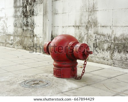 Thai red fire hydrant or red street water valve, is are emergency water supply on a white concrete footpath in front of a dirty wall with stain.