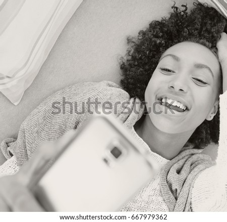 Black and white fun portrait of beautiful smiling african american young woman holding using smart phone, taking selfies pictures, networking laying on sofa, lounging. Technology recreation lifestyle.