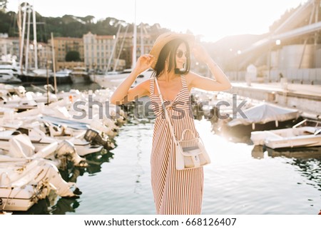 Inspired shapely girl wearing hat and little bag posing with smile in front of river in sunny day. Beautiful young woman in striped dress standing with hands up enjoying summer warm in vacation.