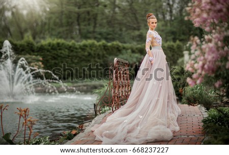 Beautiful Romantic tall Girl in fairy long lacy dress standing near pink lilac.Gorgeous young model with perfect hair style and wreath accessories dreaming in spring garden.Fantasy art work.