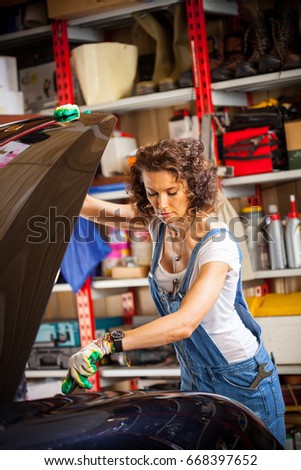 Beautiful woman automechanic in blue overall near a car with an open hood in the garage