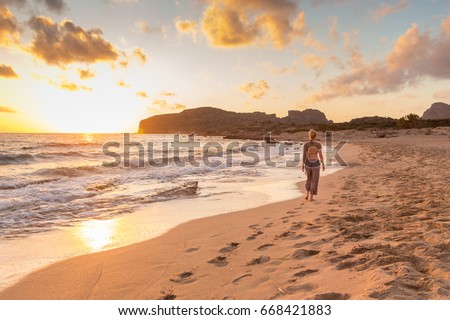 Woman walking on sandy beach at golden hour. Seashore sunset walk, Falasarna, Crete, Greece. Royalty-Free Stock Photo #668421883