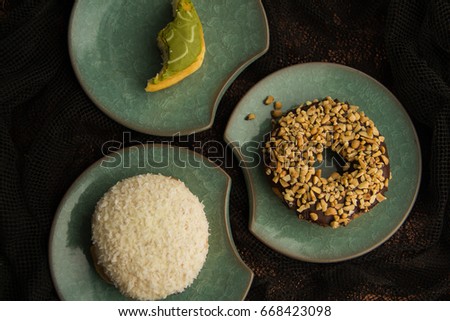 Top View Shot of Three Varieties of Donuts including a Chocolate Ring-Shaped Donut Sprinkled with Peanuts, Coconut Cake and a Bitten Matcha Filled Donut on Green Plates Dark Cloth Background