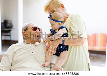 Cute little baby girl with grandmother and grandfather on wheelchair on summer day in garden. Happy seniors holding smiling child on arm.