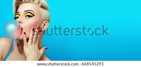 Fashionable girl with a stylish haircut inflates a chewing gum. The girl in the studio on a blue background. The girl's face with bright makeup and yellow with black shadows on the eyes. Royalty-Free Stock Photo #668545291