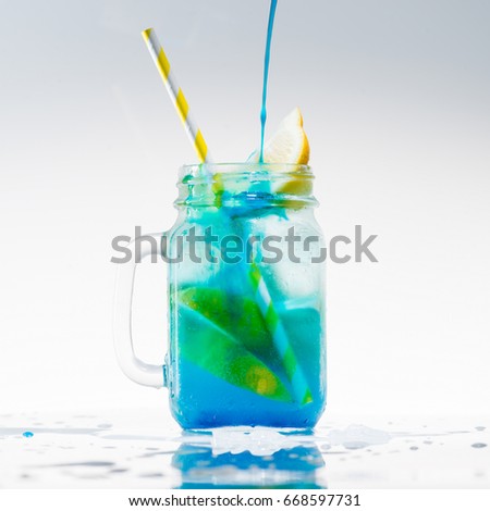 mason jar of cold blue fresh lemonade with lime, lemon and yellow drinking straw on light background. Water drops near the mason jar