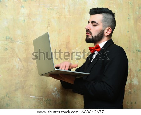 Businessman with beard, stylish haircut and busy concentrated face types on laptop doing business. Vintage beige old wall background, idea of work and leading management