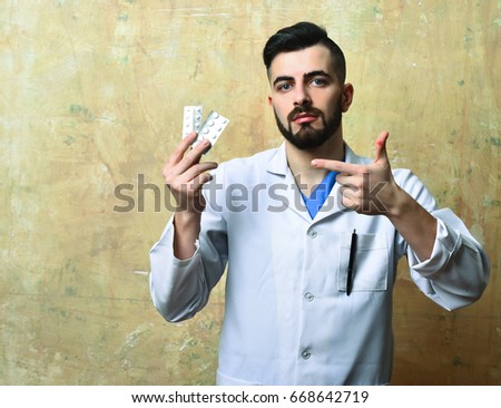 Doctor with beard and confident face expression pointing at blisters of pills on background of beige color wall. Concept of medicaments and health
