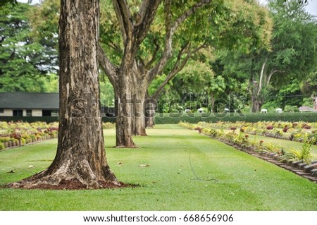 big trees and lawn in a cemetery with headstones in the background at Kanchanaburi War Cemetery in Thailand.