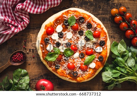 Pizza with tomatoes, mozzarella cheese, black olives and basil. Delicious italian pizza on wooden pizza board. Table top view