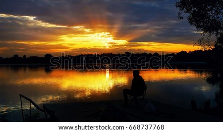 The silhouette of a seated fisherman on a beautiful background of a rising sun with rays in the dramatic clouds .
