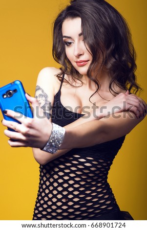 Portrait of a beautiful dark haired girl with tattoo on her arm wearing black see-through net top and jewel bracelet holding a blue cell phone and making selfie. Isolated on yellow background