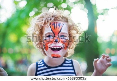smiling boy with curly hair and face art painting like tiger, little boy making face painting, halloween party, child with funny face painting, little cute boy with faceart on birthday party close up  Royalty-Free Stock Photo #668933035
