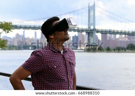 young handsome man wearing virtual reality headset goggles standing by a waterfront with a bridge and urban city skyline in the background 