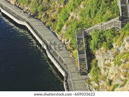 Beautiful summer season specific photograph. Pier/walkway/harbor/wooden path close to the ocean. Photo taken from above. Green vegetation, plants and rocks next to the wooden walkway.