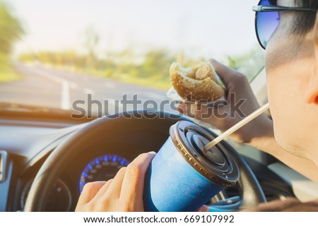 Man is dangerously eating hot dog and cold drink while driving a car Royalty-Free Stock Photo #669107992