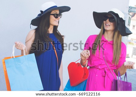 Two beautiful young cheerful women holding shopping bags and talking while looking at each other.  Beautiful girls with shopping bags standing in front of a white wall.