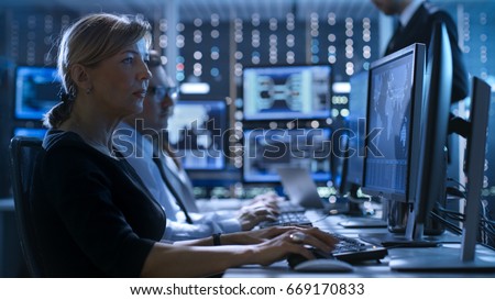 Female Government Employee Works in a Monitoring Room. In The Background Supervisor Holds Briefing. Possibly Government Agency Conducts Investigation. 