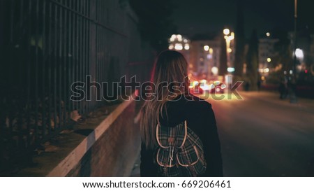 Back view of brunette woman walking near the road at the traffic time. Girl goes through the city late at night alone. Royalty-Free Stock Photo #669206461