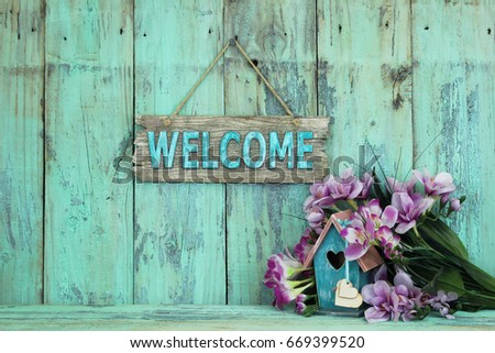 Wood welcome sign by birdhouse with heart and purple spring flowers hanging on antique rustic mint green wooden background; springtime, Easter and Mothers Day background with copy space