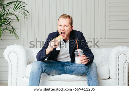 A young man in a blue jacket sits on a white couch, eats a burger and drinks a milkshake
