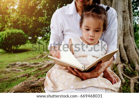 Mother and little girl reading a book under big linden tree.
