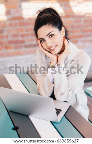 Indoor portrait of young successful Caucasian woman blogger writing a new post for her popular blog using notebook while sitting at desk over brick wall background. Smiling pretty lady with red nails