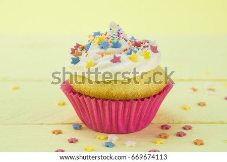 Single vanilla sponge birthday cupcake frosted and coated with star candy sprinkles on pale yellow background