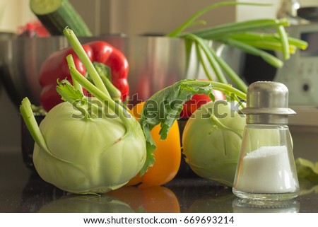 fresh green kohlrabis, red and yellow paprika, a salt shaker and other vegetables in the background of this kitchen scene. Healthy vegetables and salt shaker, placed on black worktop in the kitchen. 