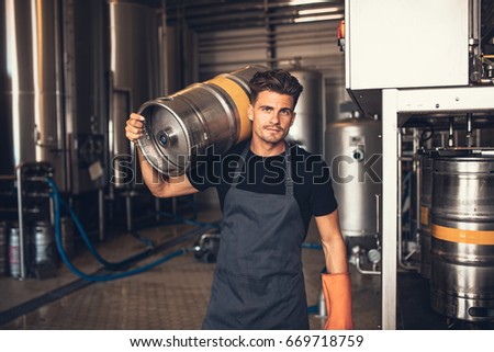 Portrait of male brewer carrying metal container at brewery factory. Young man holding a keg on shoulder at warehouse. Royalty-Free Stock Photo #669718759