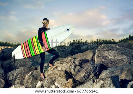Surfer with sunglasses standing on the rock and holding his surfing board with the blue sky with clouds in the background.