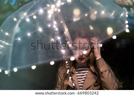 A blurry silhouette of a girl with the transparent umbrella on a city street, rain drops and a lot of lights. Rainy weather in the summer, blurred flowers, lights of Shining umbrella in raindrops