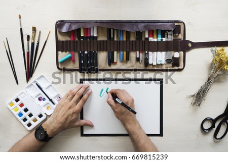 Top view on calligrapher hands writing with blue marker on blank paper on wooden table background with markers in leather case, dried yellow flowers, black scissors, brushes and watercolor palette. 