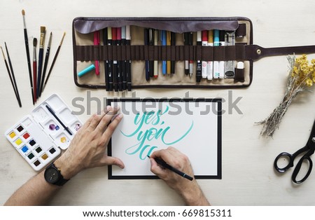 Top view on calligrapher hands writing words Yes, you can with blue marker on blank paper on wooden table background with markers in leather case, dried yellow flowers,brushes and watercolor palette. 