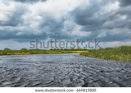 Storm clouds over river bank with reeds and trees.